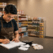 Pemilik minimarket menyusun laporan keuangan toko dengan memeriksa data pemasukan dan pengeluaran di meja administrasi.